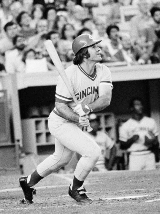 Pete Rose in a Cincinnati Reds uniform swinging at a pitch during a 1970s MLB game.